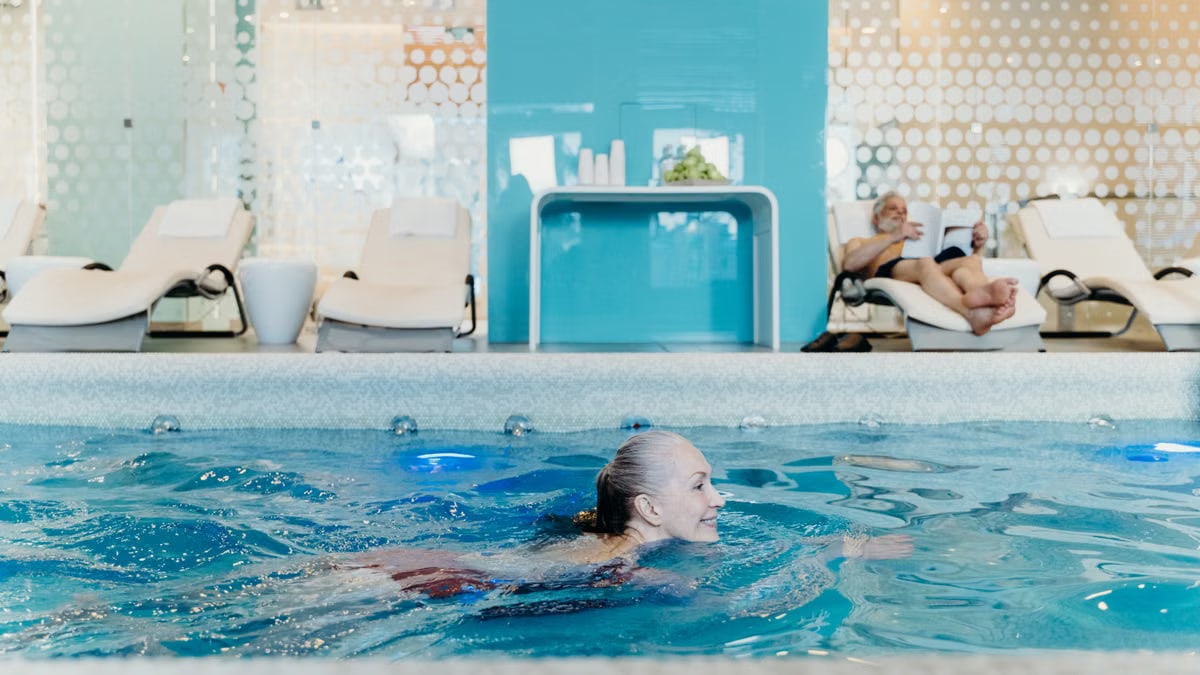 Elderly woman swimming in a modern indoor pool while another senior relaxes on a lounge chair.